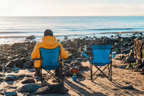 Two people on beach chairs together on winter beach, making hot tea, coffee on sunny blue sky day and the sea in background. British cold winter. Lifestyle tourism concept © Iryna