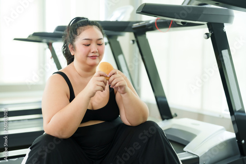 Overweight Asian woman exercise in gym, female contemplates her snack, reflecting on dietary choices in fitness journey. moment of thought, examines Hamburger, considering health and wellness goals