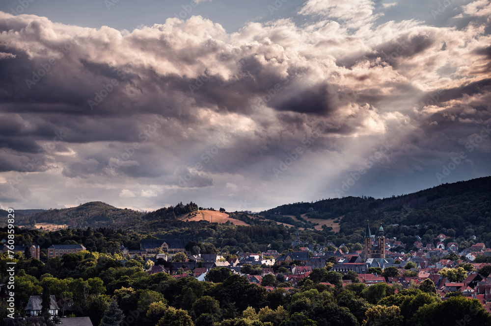 A dynamic cloudscape crowns a peaceful town, nestled among lush hills, with rays of sunlight breaking through to highlight its charm