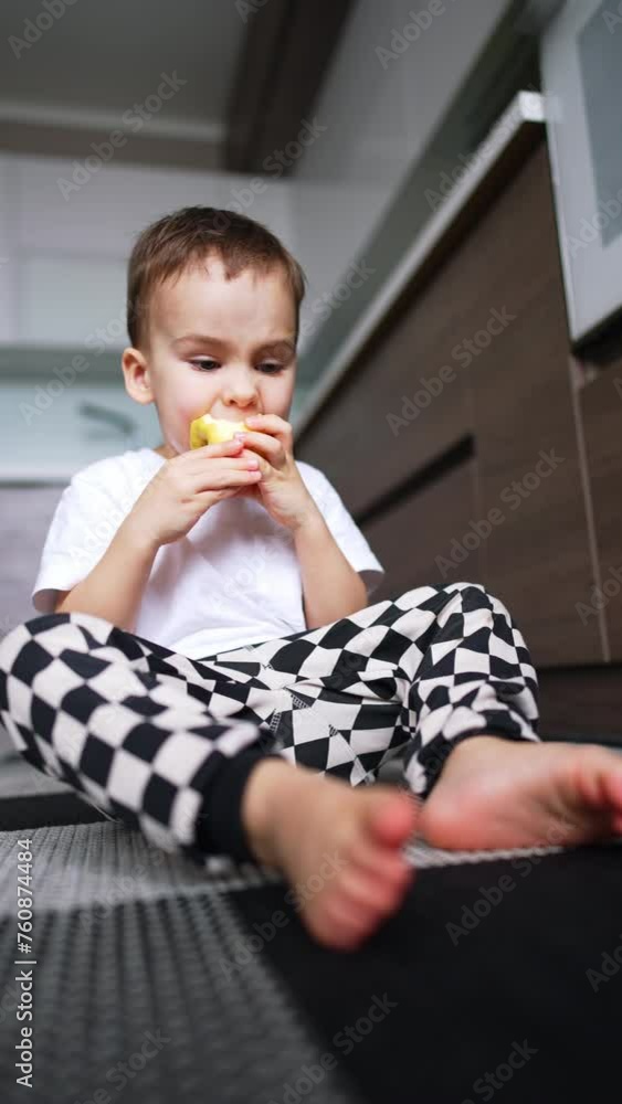 Kid in black and white checkered pants sits on the floor eating an apple. Baby boy eating fruit in the kitchen. Low angle view. Vertical video. Kid in black and white checkered pants sits on the floor eating an apple. Baby boy eating fruit in the kitchen. Low angle view. Vertical video.