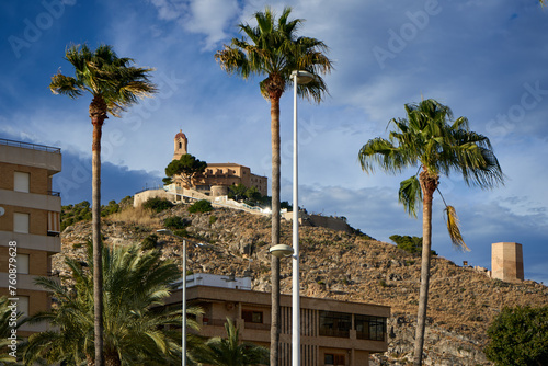 Castle of Cullera. A stunning scene unfolds with a grand building atop a hill, surrounded by palm trees and lush greenery. photo