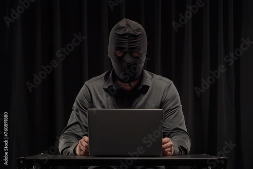 A man in a black robber mask sits with a laptop isolated on a black background
