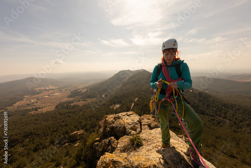 A woman is standing on a mountain top, wearing a helmet and harness. She is looking out over the landscape, taking in the view. Concept of adventure and excitement