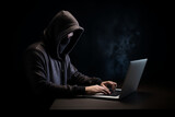 Man on dark background in a hood hiding his face working at a laptop, personal data hacking, hacker