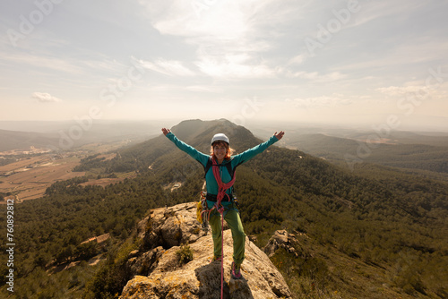 A woman is standing on a mountain top, wearing a helmet and a backpack. She is smiling and she is enjoying the view