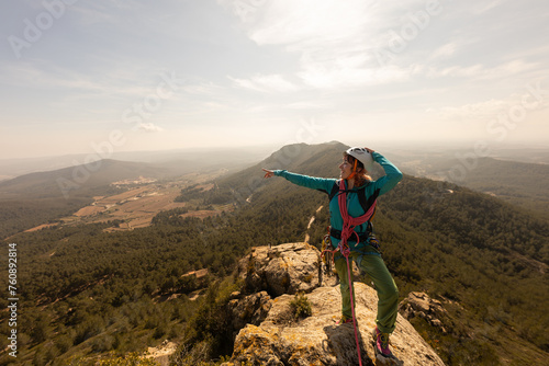 A woman is standing on a mountain top pointing to the sky. She is wearing a blue jacket and a backpack