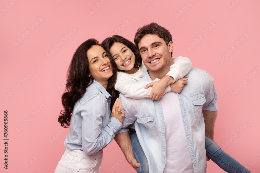 Excited family of three having fun on pink background, girl piggybacking dad, happy young parents and their daughter smiling at camera Excited family of three having fun on pink background, girl piggybacking dad, happy young parents and their daughter smiling at camera