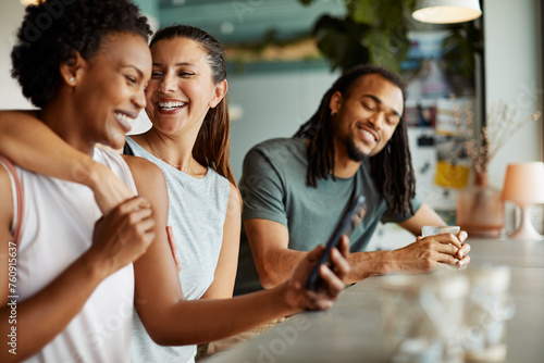 Three diverse friends laugh and fool around in a cafe after a yoga class. They are looking at funny pictures on a cell phone photo