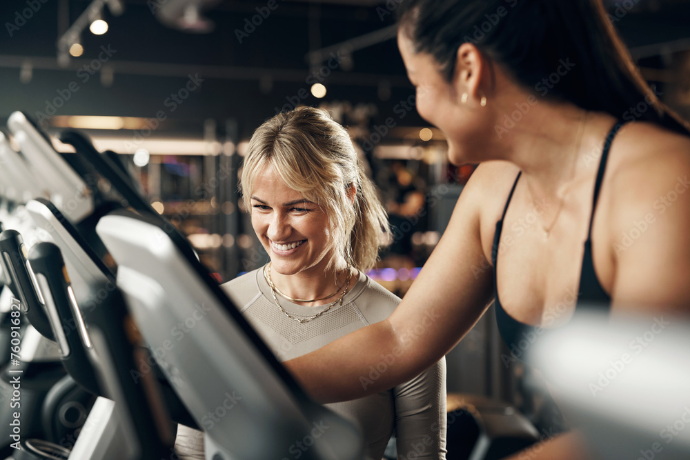Female instructor laughing while helping a fit young woman riding on a stationary bike during a cardio exercise session in a gym Female instructor laughing while helping a fit young woman riding on a stationary bike during a cardio exercise session in a gym