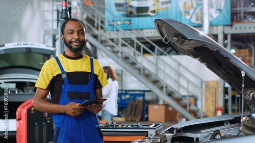 Portrait of smiling mechanic in car service ordering new parts for damaged vehicle using tablet. African american repairman looking online for components to replace old ones in busted automobile