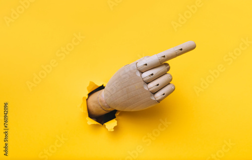 A wooden hand protrudes from a torn hole in yellow paper and points its index finger to the right. Concept of direction, adjustment and assistance. Mannequin, robot
