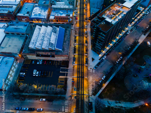 aerial drone view of Urban streets, bathed in warm twilight hues, form a symmetrical pattern. Buildings with textured rooftops flank the illuminated roads. A tranquil view from above, 
