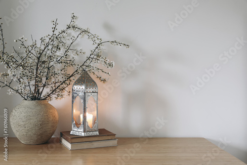White flowers, blooming prunus tree branches in vase. Glowing Moroccan lantern on books. Wooden table. White wall background. Iftar dinner. Ramadan Kareem greeting card. Eid ul Fitr muslim holiday.
