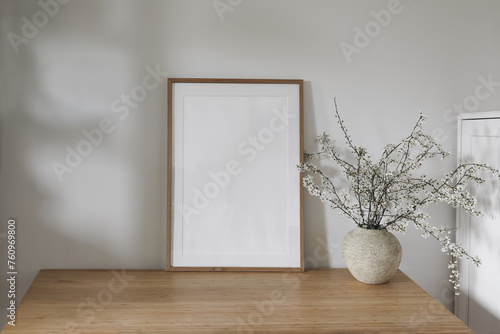 Vertical wooden picture frame, poster mockup in sunlight. Spring composition. Elegant interior, home office still life. Artistic display. Blooming cherry plum tree branches in vase. Wooden table, desk