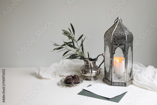 Ramadan Kareem still life. Glowing ornamental lantern. Olive tree branches in silver jug, vase.Blank vard mockup. Date fruit, silver tray. Iftar dinner. Eid mubarak, Eid ul Fitr holiday celebration.