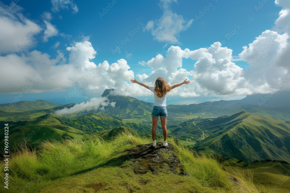 Female traveler with arms outstretched standing on a mountain summit overlooking green hills and clouds. Young woman cheering arms up of joy