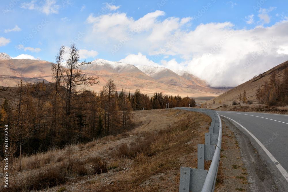 An asphalt road with metal fences descends from the mountain into a picturesque autumn valley at the foot of snow-capped mountains on a sunny autumn day. An asphalt road with metal fences descends from the mountain into a picturesque autumn valley at the foot of snow-capped mountains on a sunny autumn day.