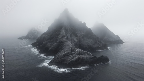 Atmospheric black and white photograph of a misty, jagged rock formation rising from the ocean, evoking mystery and the sublime power of nature