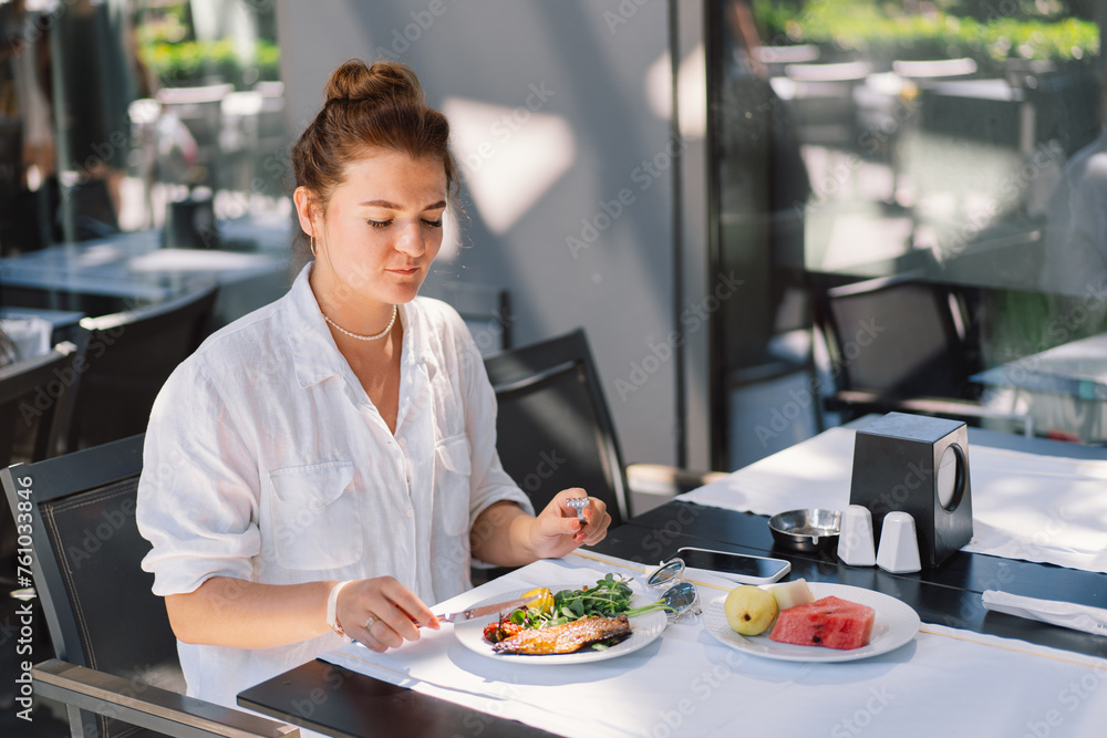 A woman in a white shirt eats lunch or breakfast outdoors in a cafe. Woman eating healthy food on a restaurant terrace A woman in a white shirt eats lunch or breakfast outdoors in a cafe. Woman eating healthy food on a restaurant terrace