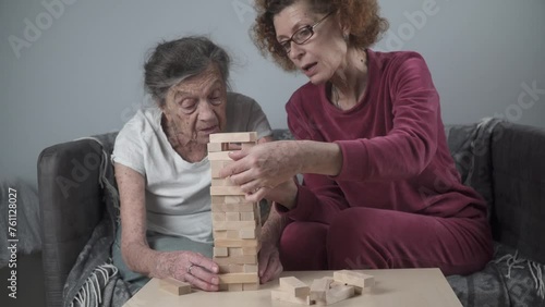 Retired mother and daughter spend time together at home, playing board game and caressing dachshund dog. Caucasian senior woman builds tower of wooden blocks with volunteer assistant in nursing home. photo