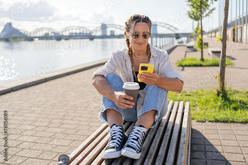 Hipster female with braids and takeaway coffee sitting using smartphone at street of city.Happy student girl sitting outside with smartphone in hands having coffee break, have video call with friends