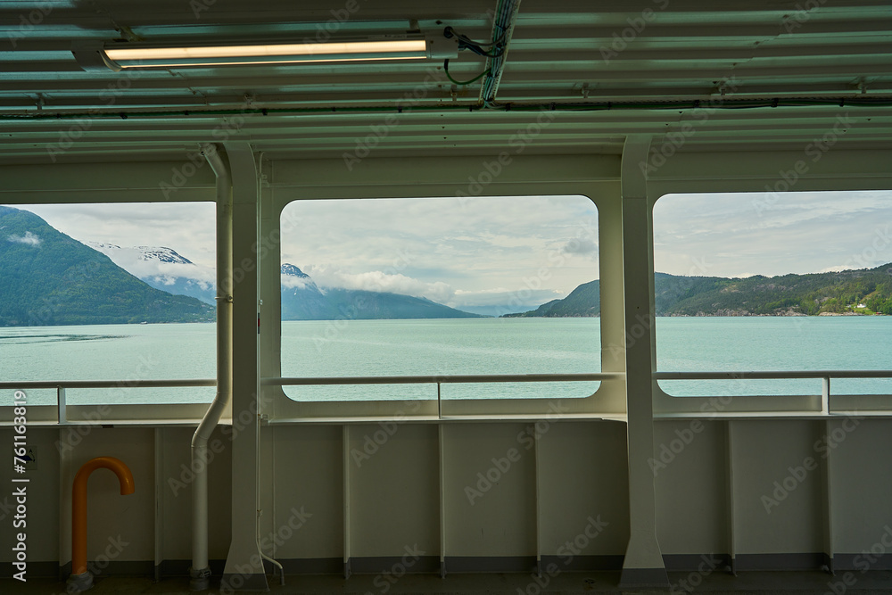 typical car ferry in Norway crossing the Hardanger Fjord with turquoise blue water on a sunny day with snow covered mountains in the background.