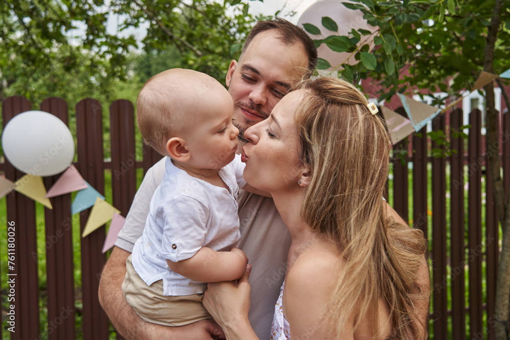 Fototapeta premium Mom kisses the baby, who is sitting in Dad's arms. happy young family surrounded by green trees and festive flags