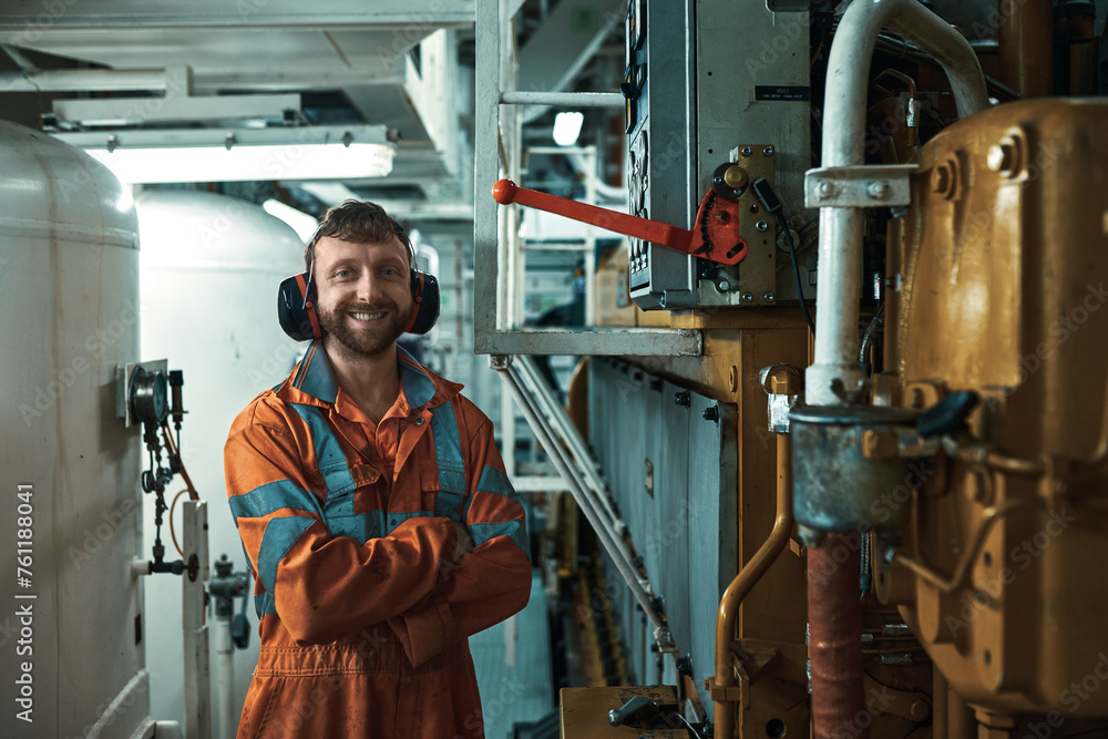 Smiling engineer in orange boiler suit, standing near main engine control box in engine room. Smiling engineer in orange boiler suit, standing near main engine control box in engine room.