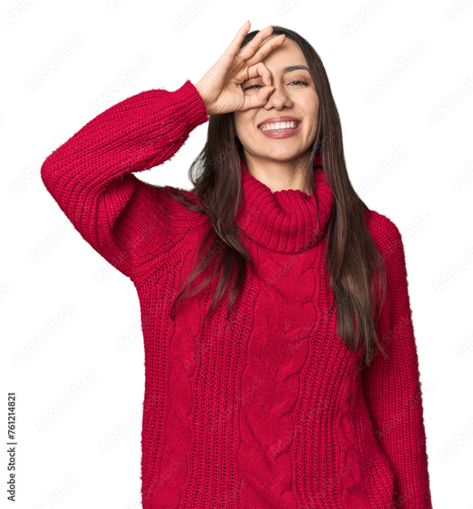 Young Caucasian woman in cozy red sweater on studio background excited keeping ok gesture on eye. Young Caucasian woman in cozy red sweater on studio background excited keeping ok gesture on eye.