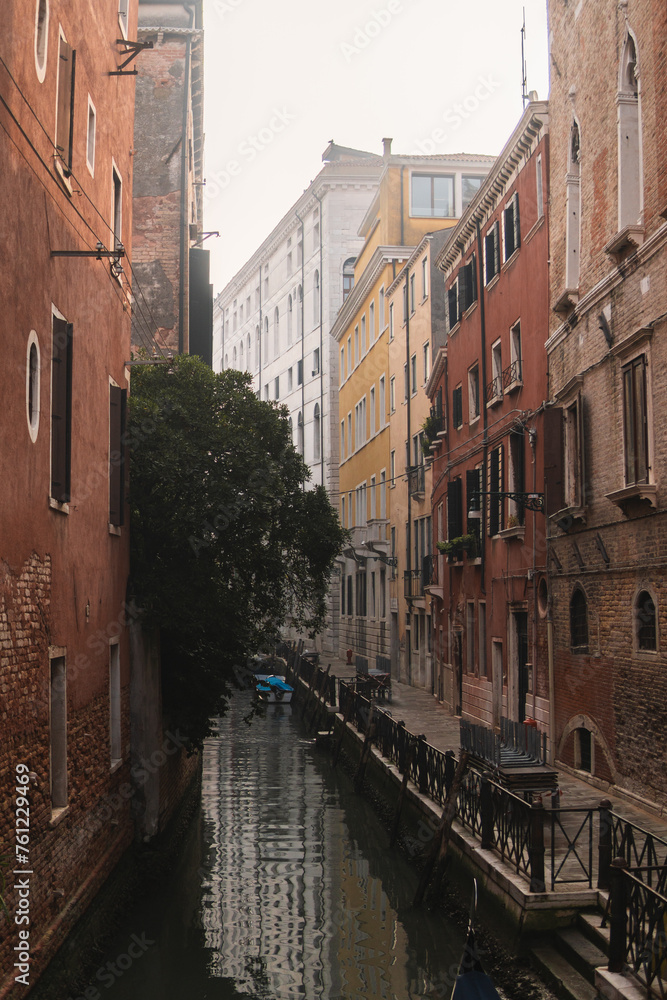 Tranquil Venetian canal scene with charming brick and colorful buildings, a lush tree leans over the calm water, where the scene is reflected, with a boat gently moored by the quay, on a cloudy day.