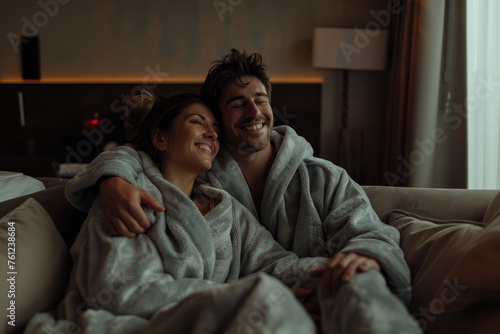 A beautiful young woman in white is sitting on the bed, smiling and looking at her husband who stands beside her with his hands around her shoulders