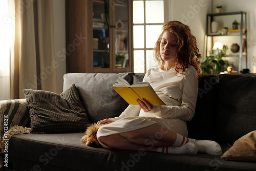 Pregnant woman reading book sitting with dog on sofa photo