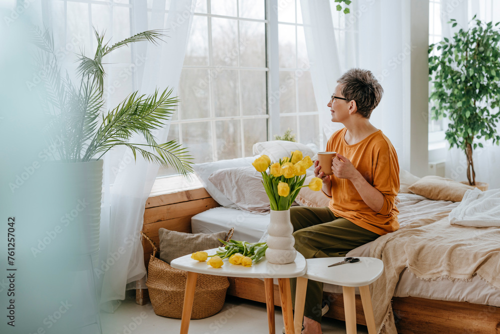 Woman sitting on bed near tulips and looking through window at home Woman sitting on bed near tulips and looking through window at home