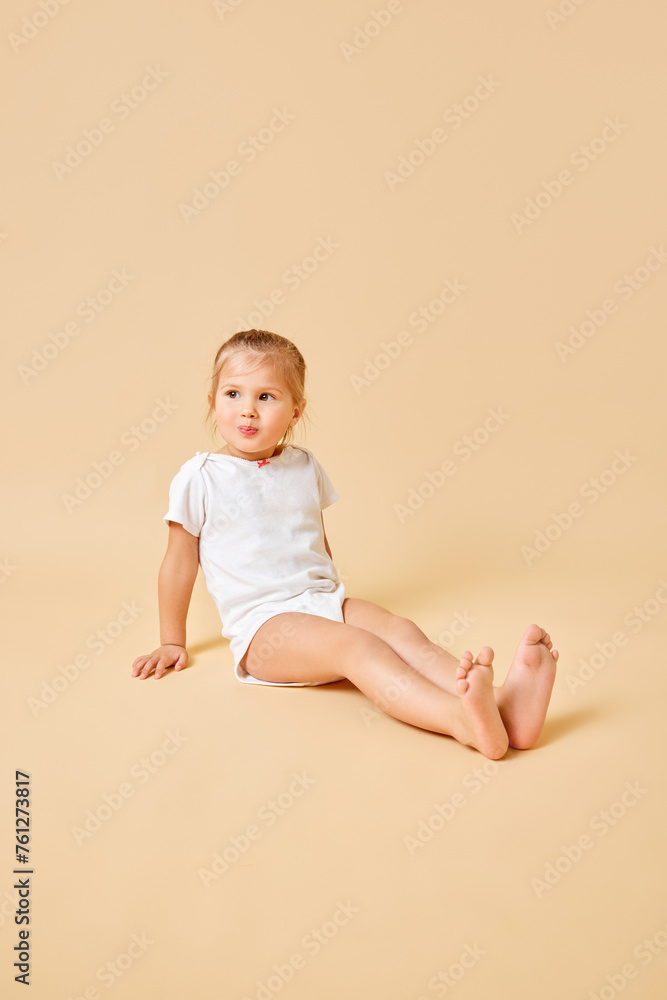Joyful little baby, pretty girl in white romper sitting on floor with cute cheerful smile against beige studio background. Concept of childhood, motherhood, life, birth. Copy space for ad Joyful little baby, pretty girl in white romper sitting on floor with cute cheerful smile against beige studio background. Concept of childhood, motherhood, life, birth. Copy space for ad