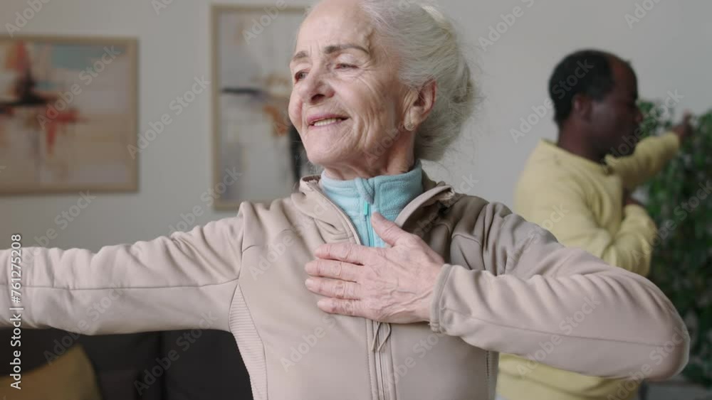 Waist up shot of cheerful Caucasian old woman with African American husband spreading their arms one by one and breathing while warming up before morning domestic fitness training Waist up shot of cheerful Caucasian old woman with African American husband spreading their arms one by one and breathing while warming up before morning domestic fitness training