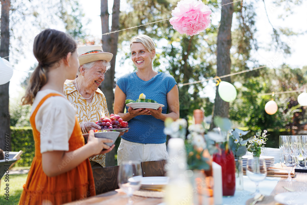 Girl helping grandmother and mother to set outdoor table for summer garden party. Bringing plates, food, and drinks. Girl helping grandmother and mother to set outdoor table for summer garden party. Bringing plates, food, and drinks.