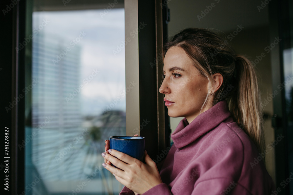 Woman enjoying cup of hot tea, coffee at home after work, standing by window wearing pink hoodie. Work-life balance for healthcare worker. Woman enjoying cup of hot tea, coffee at home after work, standing by window wearing pink hoodie. Work-life balance for healthcare worker.