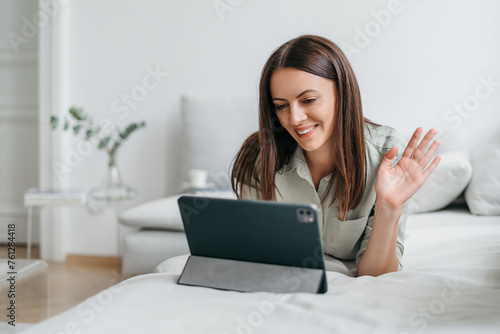 Cheerful young woman waving hand waving hand into camera with a smile. Webcam view of attractive girl sitting on the couch. Video call, virtual conference © Oleksandr