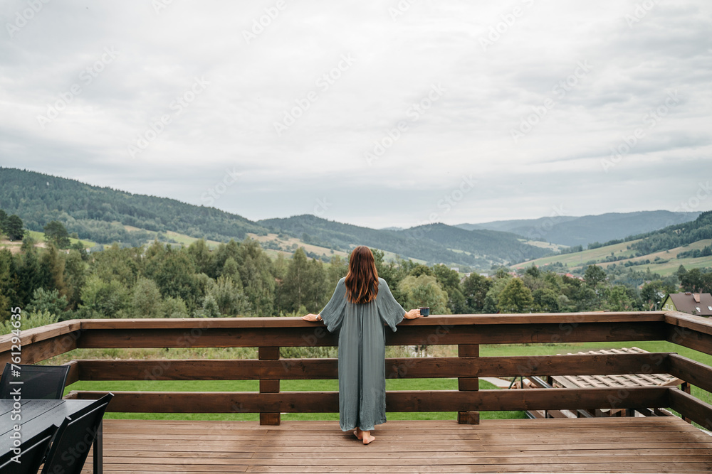 Young woman staying on terrace of wooden modern house with panoramic windows near pine forest. Concept of solitude and recreation on nature. Wellness and mindful resort. Beautiful place for vacation. Young woman staying on terrace of wooden modern house with panoramic windows near pine forest. Concept of solitude and recreation on nature. Wellness and mindful resort. Beautiful place for vacation.