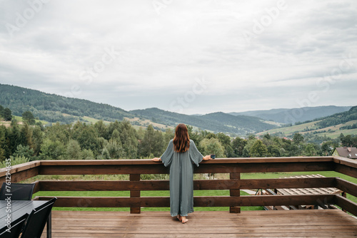 Young woman staying on terrace of wooden modern house with panoramic windows near pine forest. Concept of solitude and recreation on nature. Wellness and mindful resort. Beautiful place for vacation.