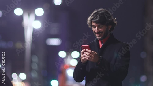 View of young man with smartphone at night time with city view landscape and traffic lights in the background. High quality video.