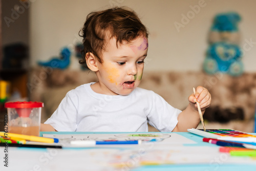 a small cute boy draws with brushes and colored paints on a sheet of paper