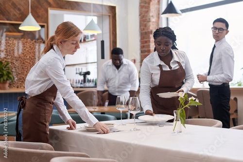 Ethnically diverse waiting staff working in modern restaurant preparing tables for guests in morning photo