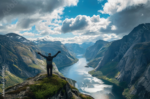 a man standing on the edge with his arms raised up, dramatic sky, overlooking a lake and mountains. A panoramic landscape of Norwegian nature © Kien