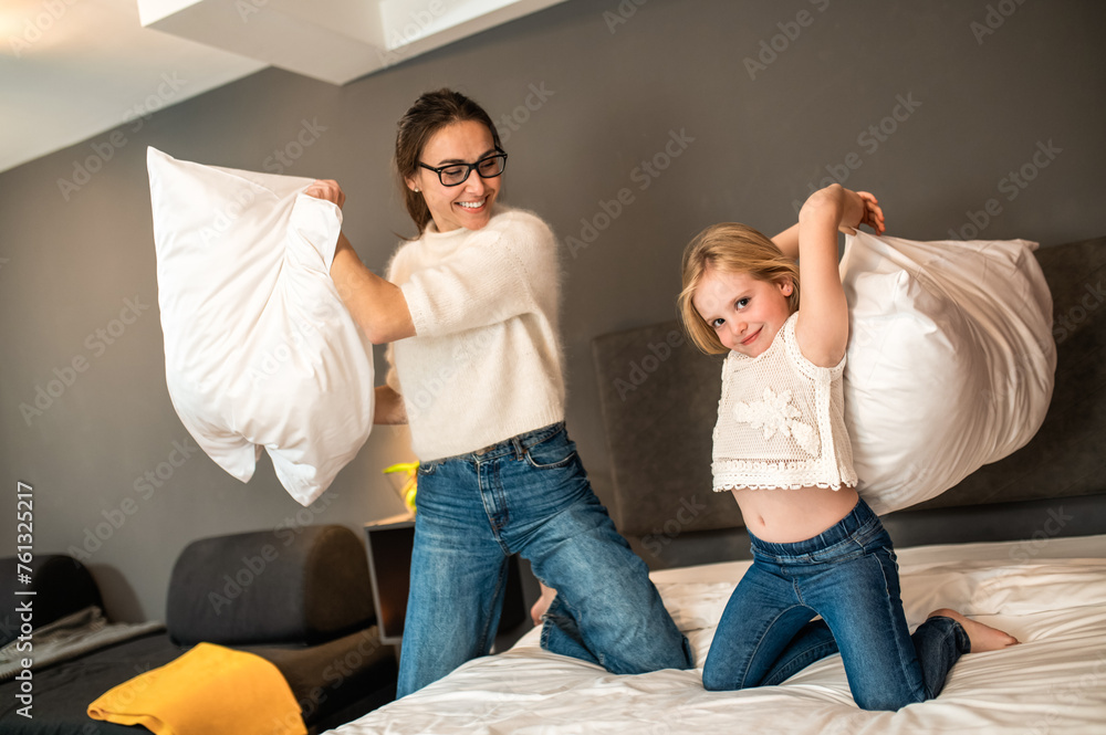 Mother and her little daughter playing with cushions on bed in hotel room - obrazy, fototapety, plakaty 