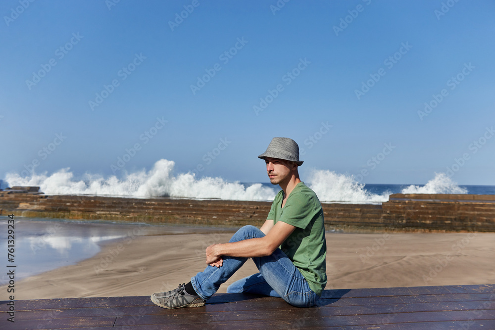 Man in casual attire rests on a dock, gazing at the sea, with waves crashing in the background. Tenerife, Spain