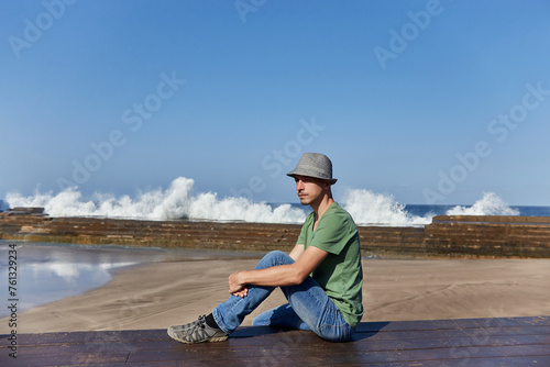 Man in casual attire rests on a dock, gazing at the sea, with waves crashing in the background. Tenerife, Spain