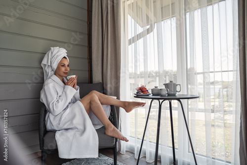 A young girl sits in her robe by the window drinking coffee. A woman in hotel in white robe, resting. Beautiful girl with a towel on her head and robe, in a hotel in an armchair relaxing on vacation.