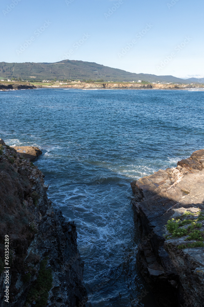 A scenic view of a rocky shoreline with waves crashing, overlooking a vast blue ocean, backed by distant green hills