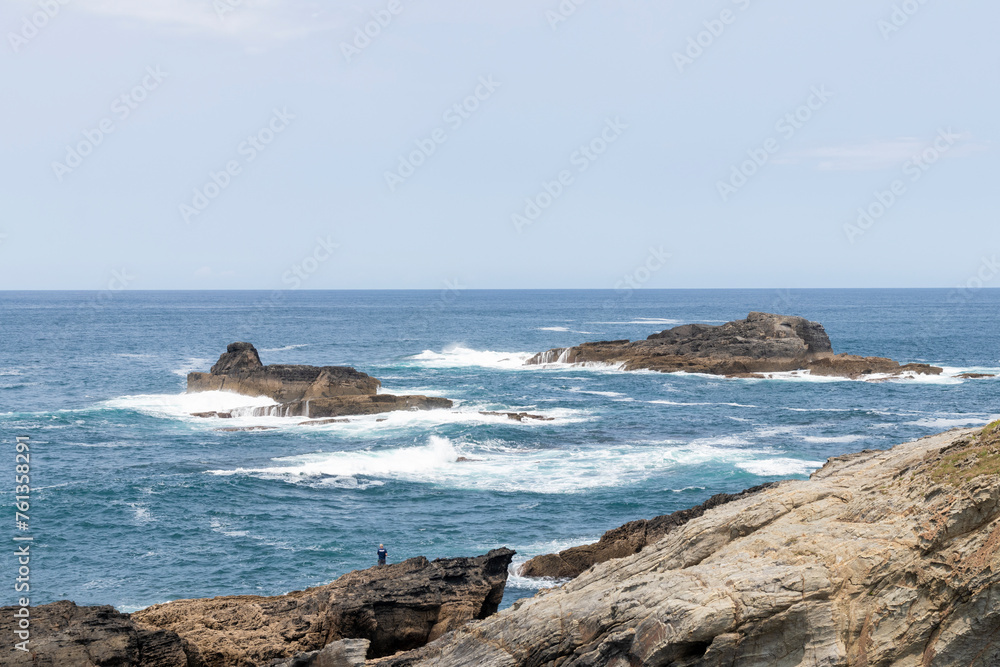 Rocky shores with powerful waves crashing, a person observing the vast, beautiful ocean and sky, evoking solitude and awe.